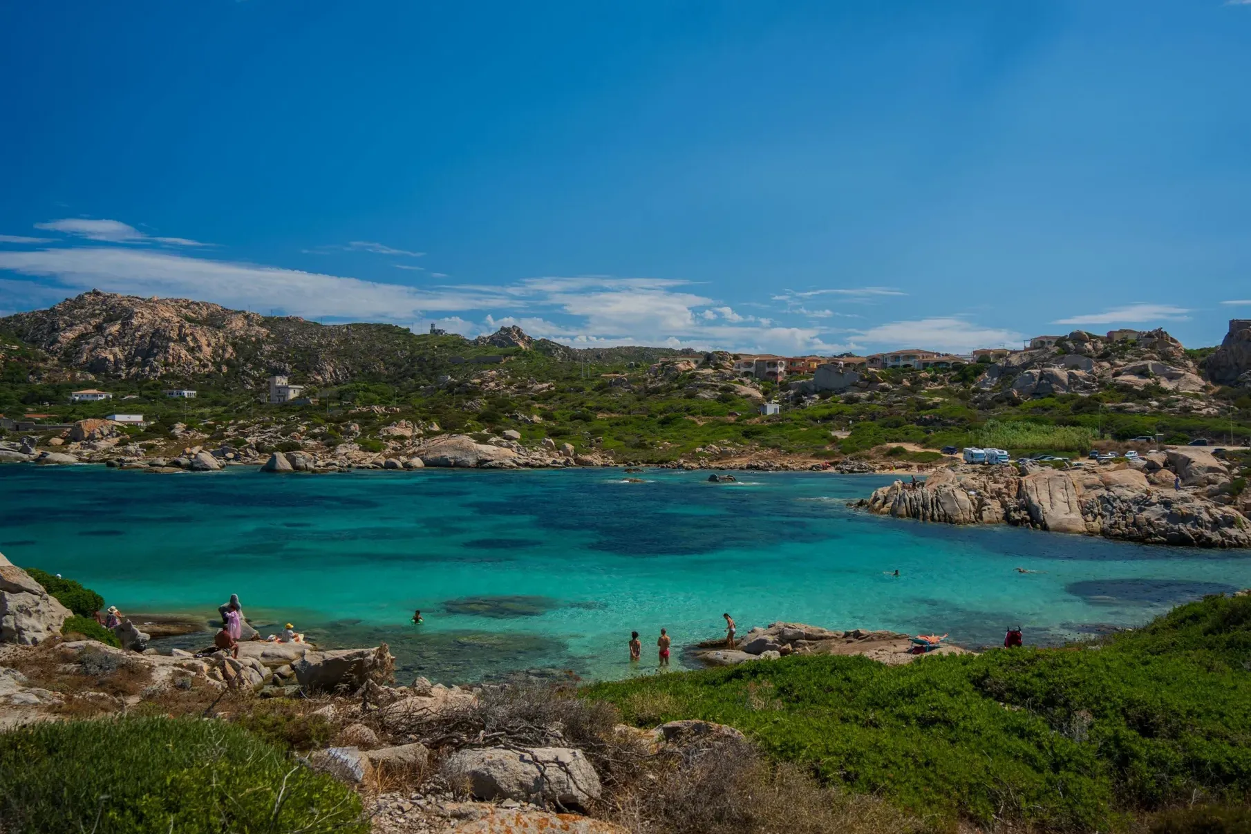 Une baie paisible aux eaux turquoise est entourée de collines rocheuses et d’arbustes verdoyants. Des personnes profitent de la baignade et de moments de détente sous un ciel bleu éclatant.