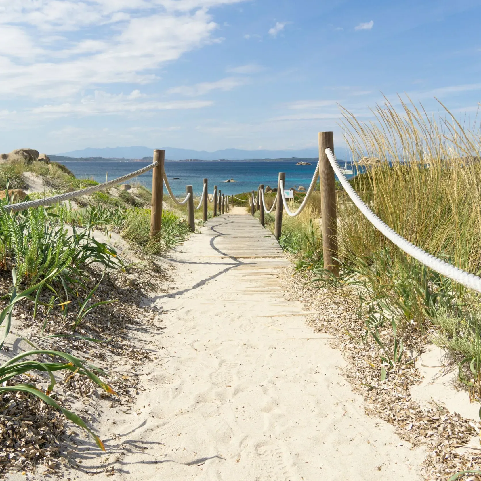 Un sentier sablonneux bordé de poteaux en bois et de cordes traverse des dunes herbeuses jusqu’à une mer bleue et calme sous un ciel dégagé, évoquant une scène de plage sereine et accueillante.