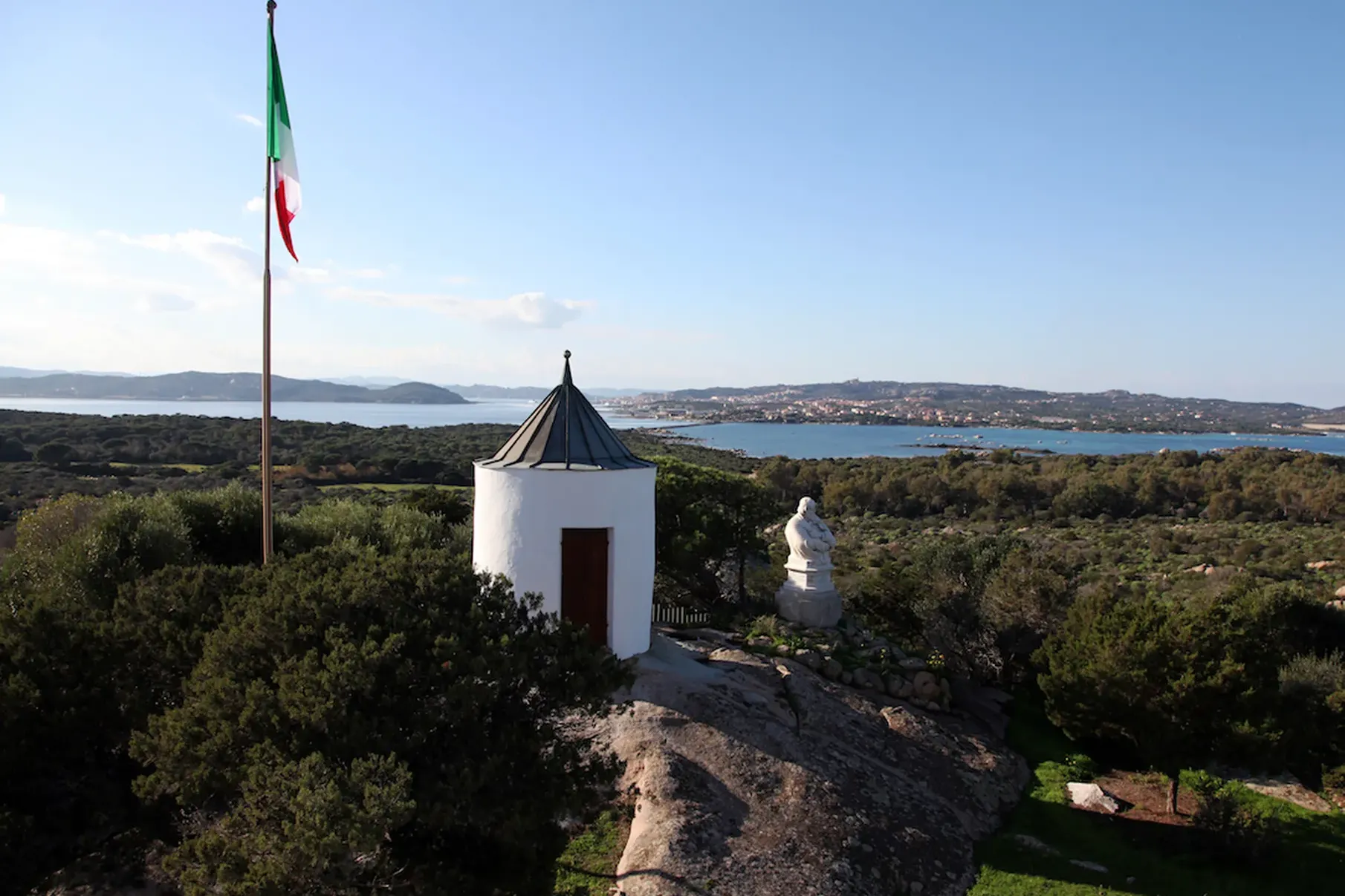 Vue panoramique d’un petit bâtiment blanc au toit conique et à la porte marron, à côté d’un drapeau italien sur une colline rocheuse. Il surplombe une baie et une ville au loin.