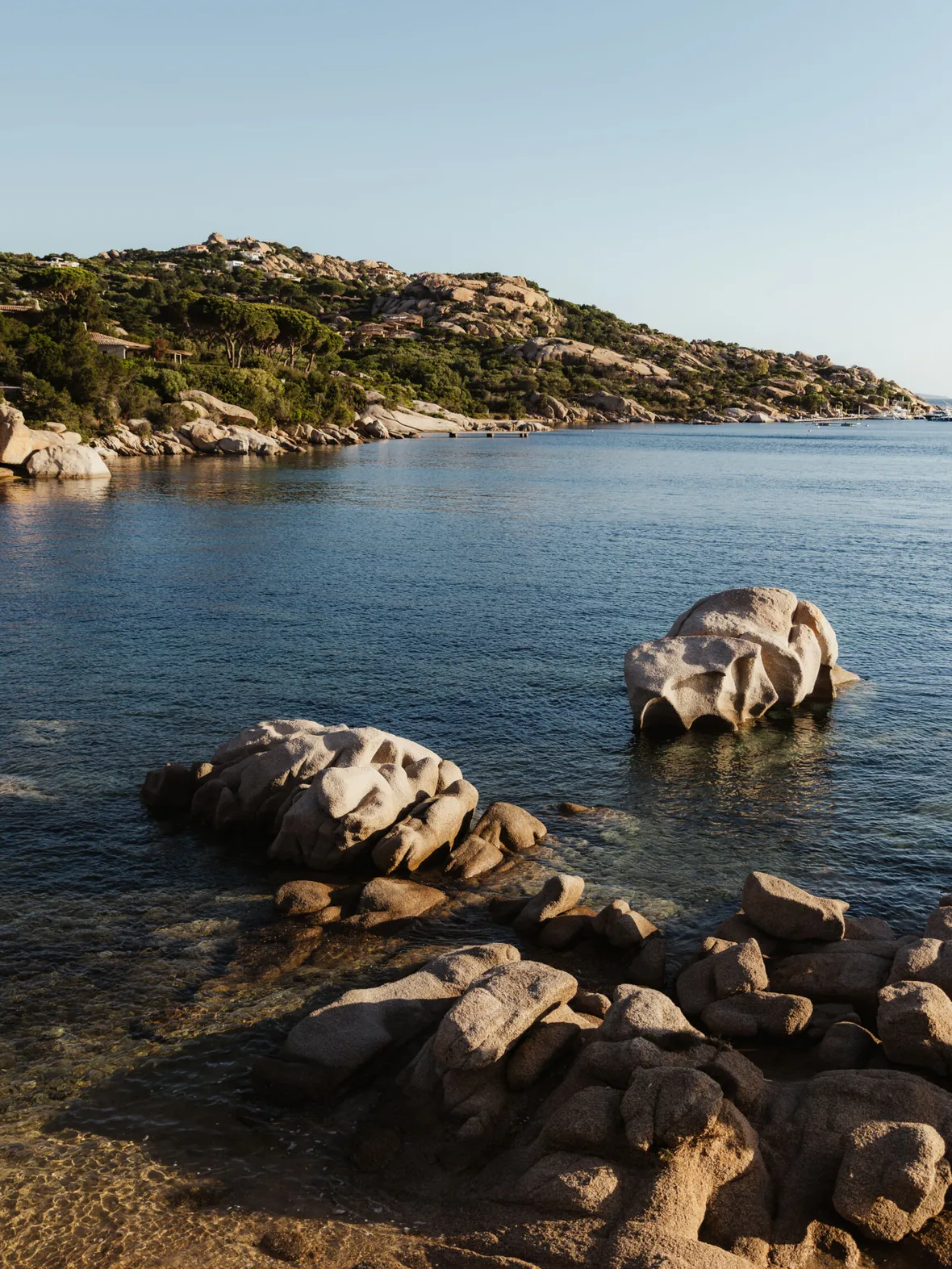 Coastal scene with smooth, sunlit rocks in clear blue water, backed by tree-lined hills under a clear sky; conveys serenity and natural beauty.