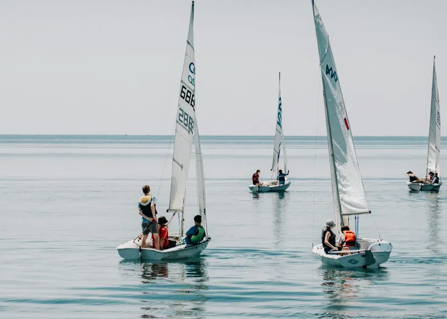Several small sailboats with people wearing life jackets are sailing on calm, open water under a clear sky during a bright, sunny day at sea.