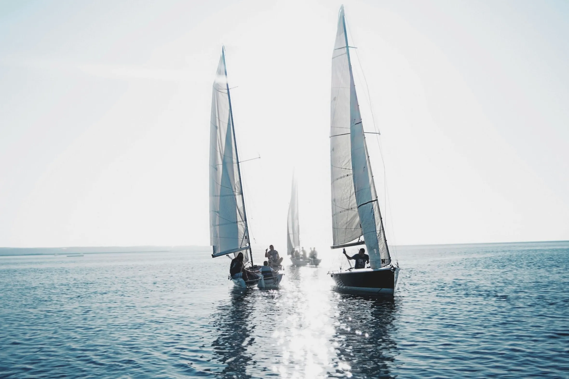 Several sailboats with white sails navigate calm blue water under a bright, hazy sky, with people visible on the decks of the vessels in the sun.