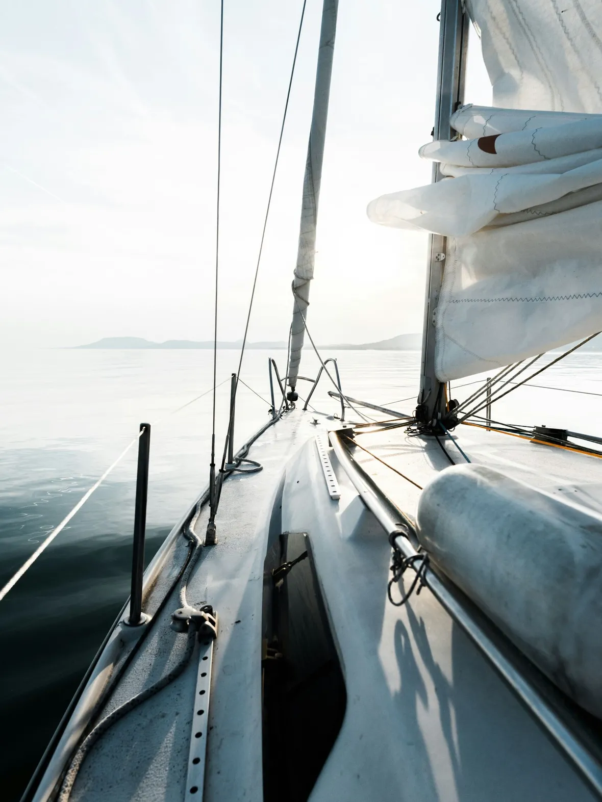 A view from the deck of a sailboat looking toward the bow, with white sails, rigging, and a calm sea under a bright, hazy sky in the distance.