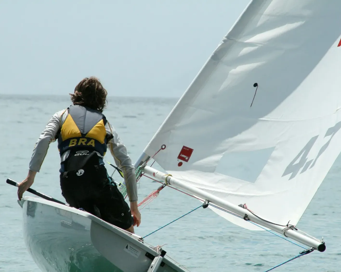 A sailor in a life vest labeled BRA sits in a small white sailboat on the water, steering with a tiller while the white sail catches the wind.