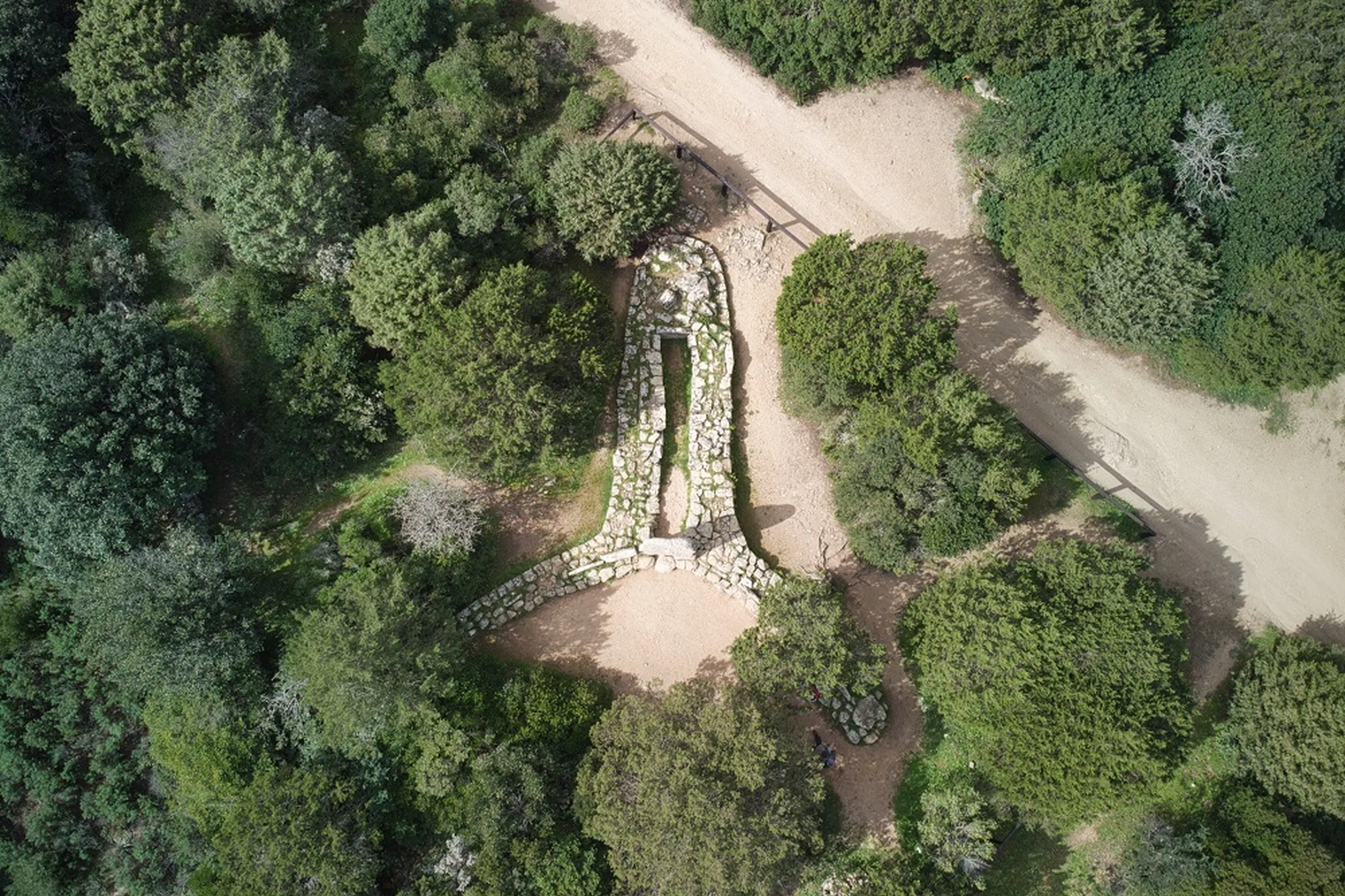 Aerial view of a stone structure shaped like a keyhole surrounded by lush green trees. A dirt path runs alongside, creating a serene, natural setting.