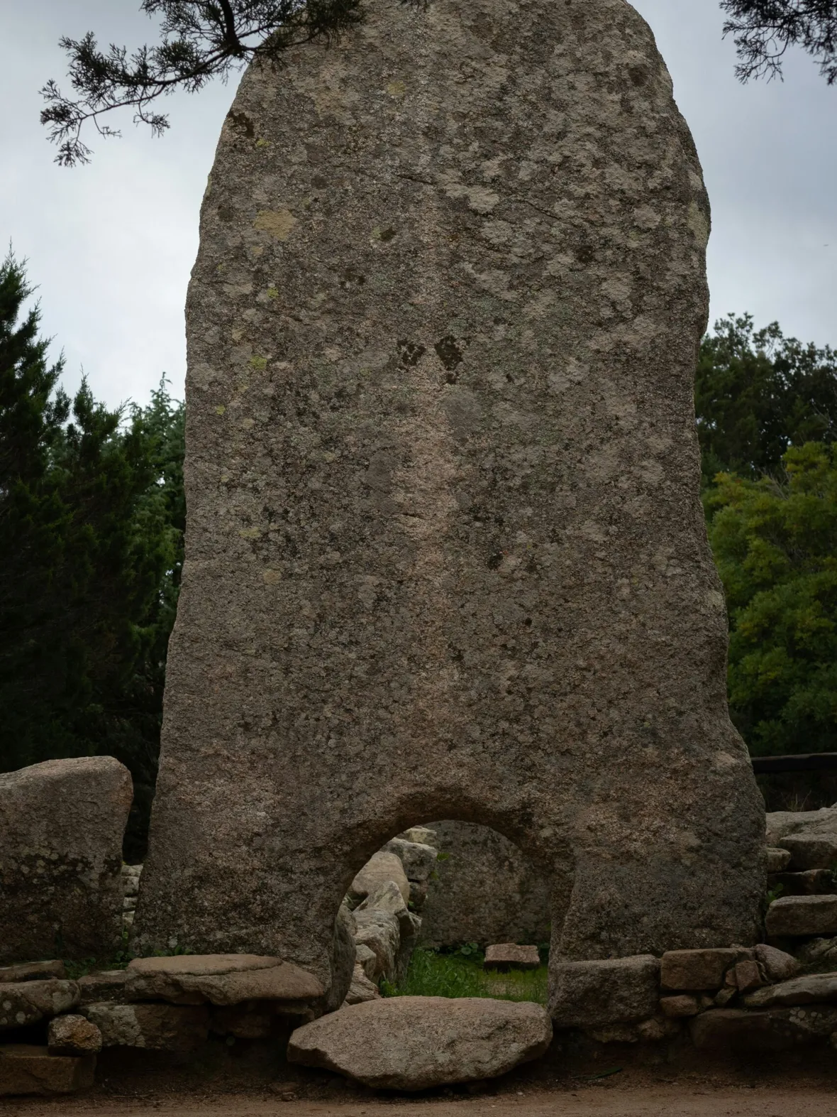 Ancient stone monolith with a weathered surface and a small archway at the bottom. Surrounded by lush trees, conveying a historical and mysterious atmosphere.