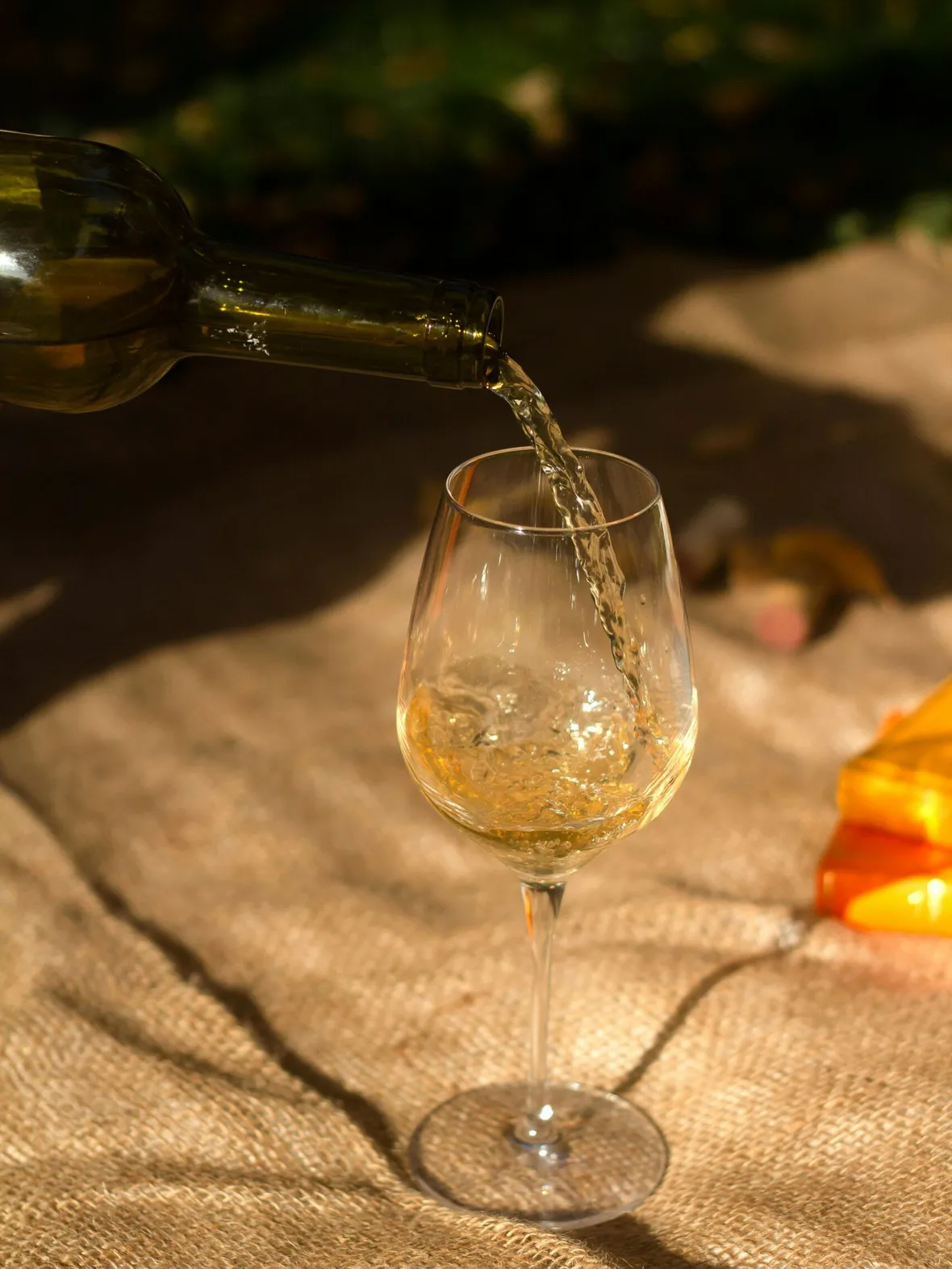 A warm-toned image of white wine poured into a glass on a burlap surface outdoors, capturing a serene, sunlit autumn ambiance with soft shadows.