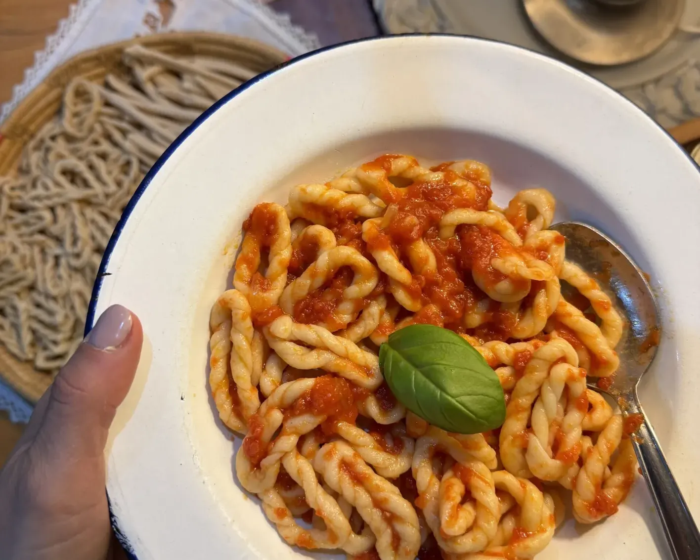 A bowl of twisty pasta with vibrant red tomato sauce is garnished with a fresh basil leaf. A spoon rests on the side. Cozy, inviting setting.