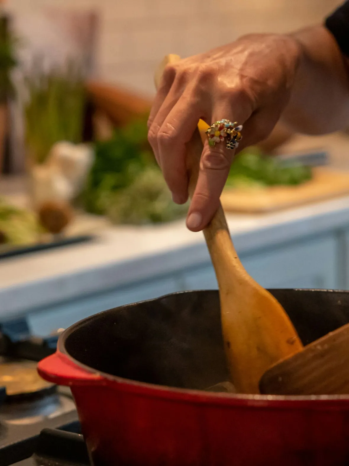 Close-up of a hand stirring contents in a steaming red pot with a wooden spoon on a stove, surrounded by herbs and vegetables on a kitchen counter.