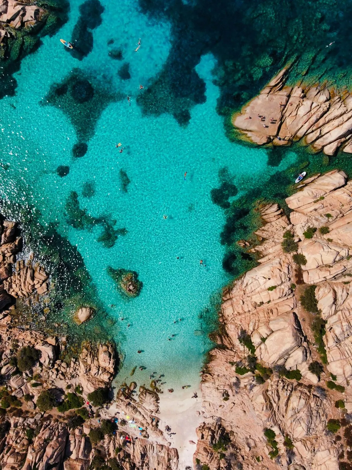 Aerial view of a rocky coastline with clear turquoise water, dotted by swimmers. Boats float near the top, conveying a serene, summer vibe.