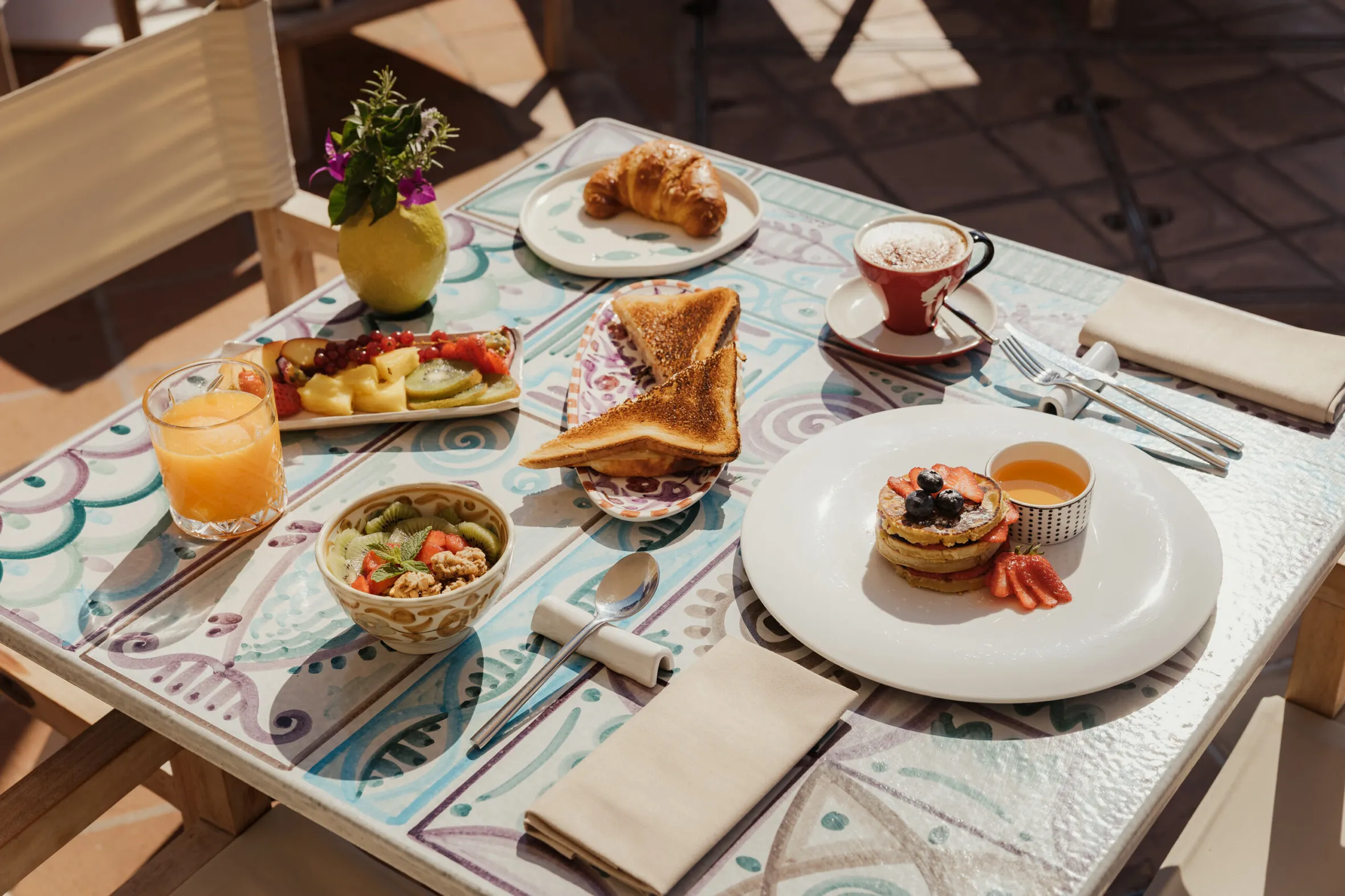 A colorful breakfast scene on a patterned table with pancakes, fruit, toast, croissant, granola, juice, and a cappuccino, evoking a sunny morning ambiance.