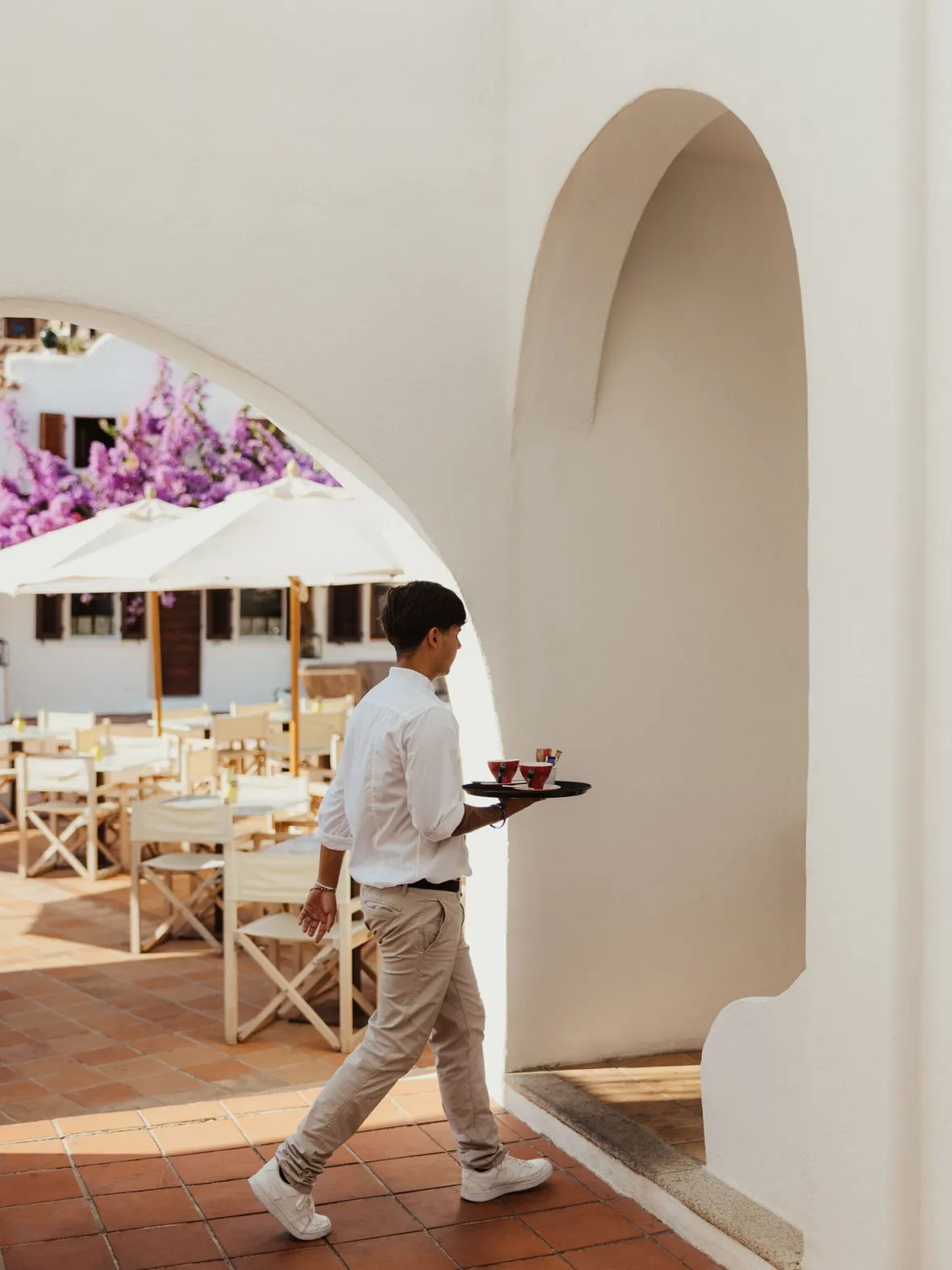 A waiter in a white shirt and beige pants carries a tray with drinks through a white archway, with outdoor tables and vibrant purple flowers in the background.